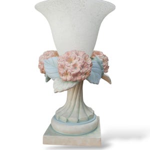 Puede incluir: Un jarrón decorativo blanco con una superficie texturizada y una parte superior acampanada. El jarrón está adornado con flores de hortensia rosas y hojas de color azul claro. La base es cuadrada y está pintada en suaves colores pastel, creando un aspecto clásico y elegante.