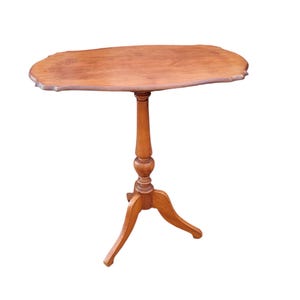 Op de afbeelding: Een lichtbruine bijzettafel in antieke stijl. De tafel heeft een geschulpte ovale bovenkant en een gedraaide voet die wordt ondersteund door drie gebogen poten. De tafel is gemaakt van hout en heeft een warme, natuurlijke afwerking.