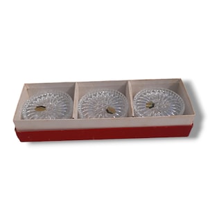 Op de afbeelding: Drie transparante glazen onderzetters met een diamantpatroon worden getoond in een rood-witte doos. De onderzetters staan in een rij en hebben elk een klein gouden label.