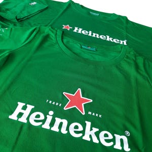 May include: Green t-shirts with the Heineken logo. The logo features the word "Heineken" in white, a red star, and the words "Trade Mark". The shirts are made of a soft material.