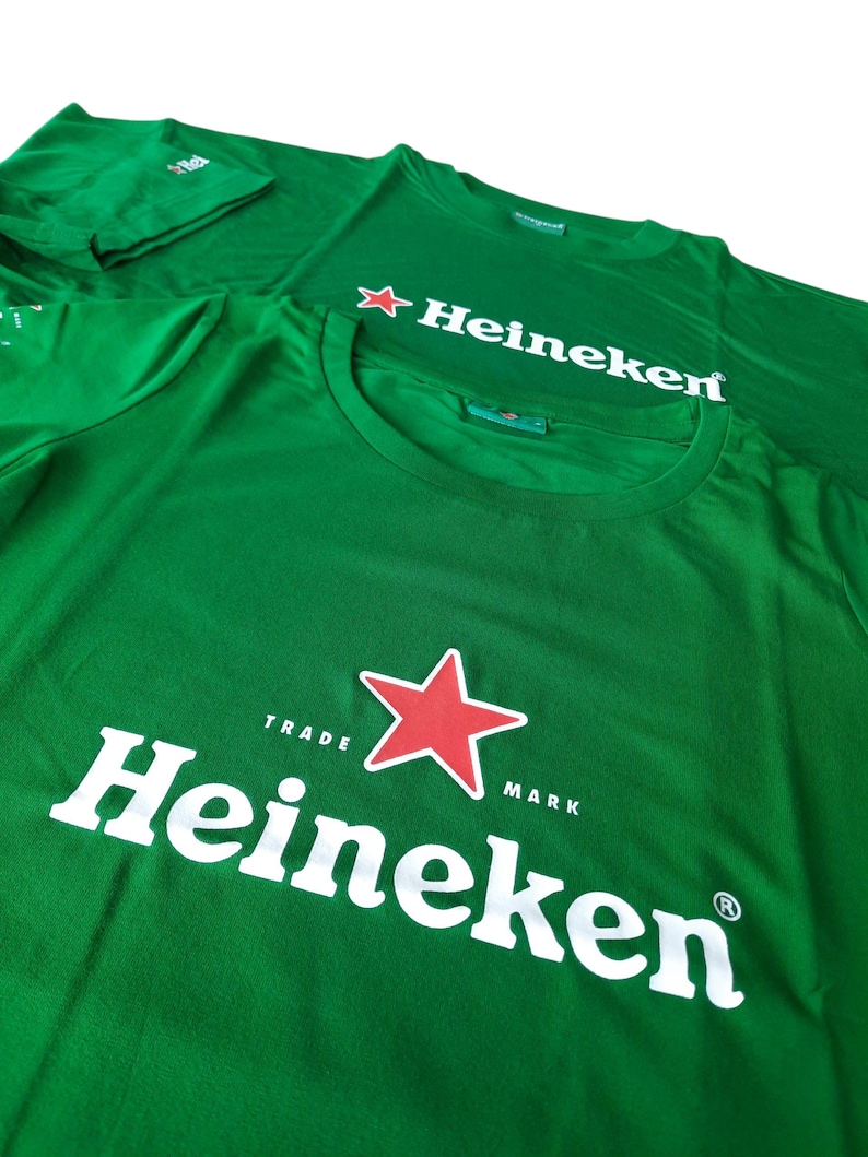 May include: Green t-shirts with the Heineken logo. The logo features the word "Heineken" in white, a red star, and the words "Trade Mark". The shirts are made of a soft material.