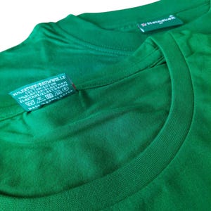 May include: Close-up of two green t-shirts. One shirt's tag reads "KATOEN/BAUMWOLLE COTTON/COTON 5% ELASTAN/ELASTHAN ELASTANE/ELASTHANNE MADE IN BANGLADESH". The other shirt's tag reads "Heineken". The shirts are made of a soft, comfortable material.