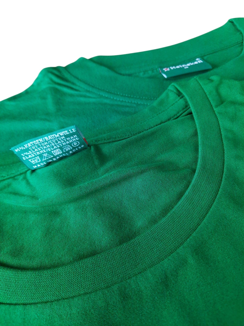 May include: Close-up of two green t-shirts. One shirt's tag reads "KATOEN/BAUMWOLLE COTTON/COTON 5% ELASTAN/ELASTHAN ELASTANE/ELASTHANNE MADE IN BANGLADESH". The other shirt's tag reads "Heineken". The shirts are made of a soft, comfortable material.