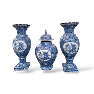 May include: A set of three blue and white ceramic vessels consisting of two tall vases with scalloped rims and square bases, and one central lidded jar. Each piece features a dense floral pattern and a central oval medallion with floral motifs.