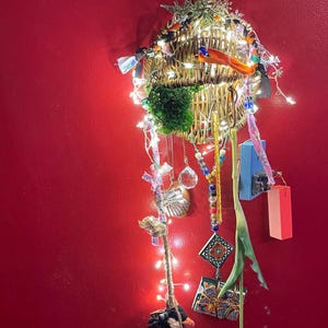 May include: A decorative wall hanging with a woven basket base, illuminated by fairy lights and decorated with ribbons and ornaments. Includes a green moss ball, crystal pendants, and a yellow tulip. Against a red wall.