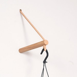 Puede incluir: Un colgador de plantas de pared de madera con una correa de cuero y un gancho de metal negro. El colgador está diseñado para sostener una planta colgante.