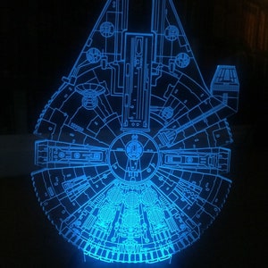 Könnte beinhalten: Ein blaues, leuchtendes Modell des Raumschiffs Millennium Falcon aus Star Wars. Das Modell besteht aus klarem Acryl und zeigt eine detaillierte Umrisszeichnung des Äußeren des Schiffes.