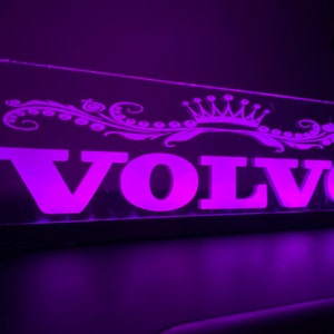 Puede incluir: Un letrero iluminado morado con la palabra "VOLVO" en letras blancas y una corona decorativa y un diseño floral sobre la palabra.