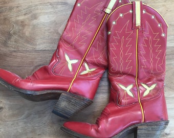 Può includere: Un paio di stivali da cowboy in pelle rossa con cuciture gialle e punta a punta. Gli stivali hanno un tacco alto e un design decorativo sullo stivale.