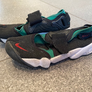 Pu&ograve; includere: Un paio di sandali Nike neri e verdi con un logo Swoosh rosso. I sandali hanno una chiusura a velcro e una suola bianca.