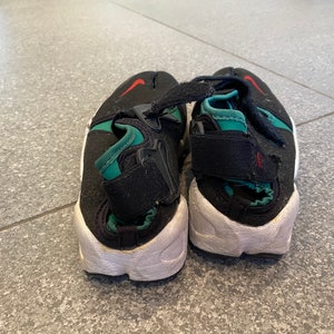 Pu&ograve; includere: Un paio di scarpe Nike nere e verdi con suole bianche. Le scarpe hanno un cinturino sulla parte superiore e uno swoosh Nike rosso sul lato.