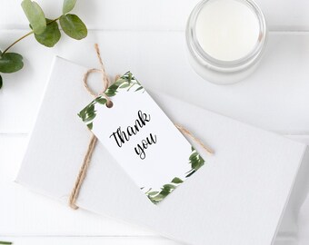 Peut inclure: Une boîte cadeau blanche avec un nœud en ficelle brune et une étiquette cadeau blanche avec un feuillage vert et le texte "Merci". La boîte est entourée de fleurs blanches, de feuilles vertes et d'une bobine de ruban blanc.
