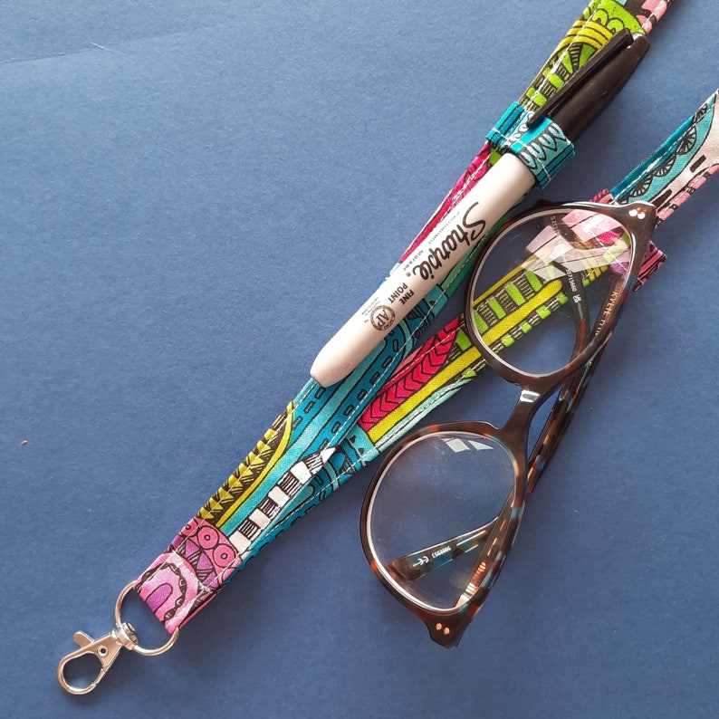 A fun fabric lanyard with a colourful funky rainbow design. A camper van appears occasionally across the print. The lanyard has a loop on each strap with a pair of glasses held in one loop and a pen in the other.