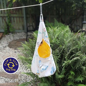 May include: A white peg bag with a floral and butterfly pattern, suspended from a metal clip. The bag has a yellow interior and the text "Flip & Clip Peg Bag" on a blue circle.