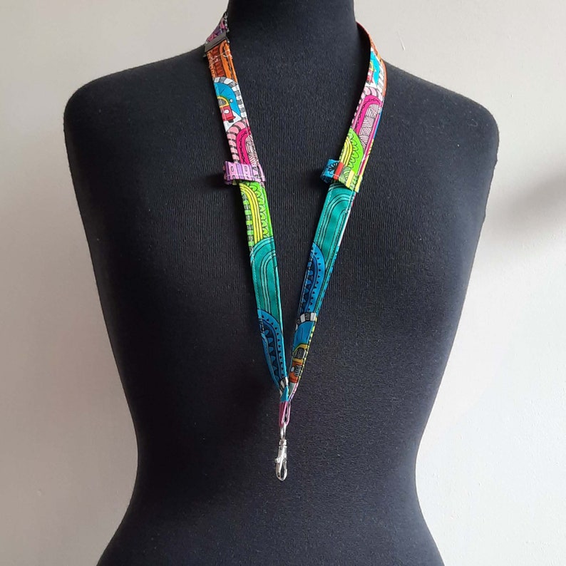 A fun fabric lanyard with a colourful funky rainbow design. A camper van appears occasionally across the print. The lanyard has a loop on each strap with a pair of glasses held in one loop and a pen in the other.