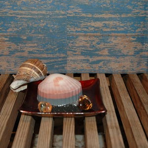 May include: A red, white, and gray layered soap bar shaped like a seashell on a red tray with two clear glass beads. The tray is on a wooden surface.