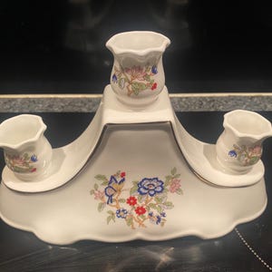 May include: A white ceramic centerpiece with three small vases, each decorated with floral patterns in pink, blue, and green. The centerpiece has a scalloped base and gold trim, with a butterfly and floral design.