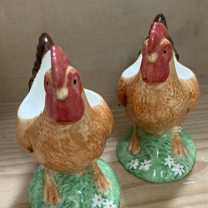 May include: Two ceramic chicken ornaments. The chickens have brown and tan bodies, red combs, and white accents. They stand on green bases decorated with white daisies. The ornaments are displayed on a wooden surface.
