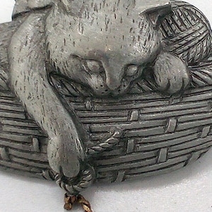 May include: A pewter cat figurine in a woven basket with a ball of yarn. The cat is curled up, with its paw extended. The basket has a rope-like border and a textured weave. A small gold-coloured chain hangs from the basket.