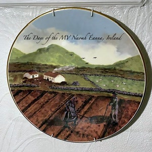 May include: A round ceramic plate depicting a painted scene of a rural Irish landscape. The plate features two figures in the foreground, a farmhouse in the middle ground, and rolling hills in the background. The text "The Days of the MV Naomh Éanna, Ireland" is printed on the plate.