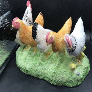 May include: A ceramic figurine of four chickens, two brown, one white with black spots, and one black and white, standing on a green grassy base.