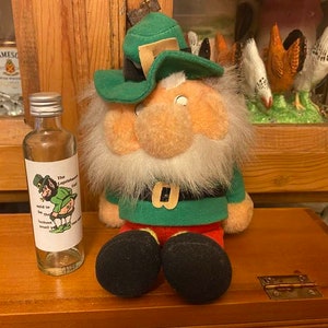 May include: A small clear glass bottle with a green and white label that says "The Leprechaun's Pot of Gold" and a cartoon leprechaun holding a pot of gold. The bottle is sitting on a wooden surface next to a green and red plush leprechaun doll.