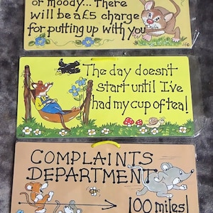 May include: Three wooden signs with humorous sayings and cartoon animal illustrations. The first sign says "If you are Irritable or moody... there will be a £5 charge for putting up with you". The second sign says "The day doesn't start until I've had my cup of tea!" The third sign says "Complaints Department > 100 miles!"