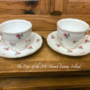 May include: Two white teacups and saucers with a pink rose pattern. The cups and saucers have a gold trim.