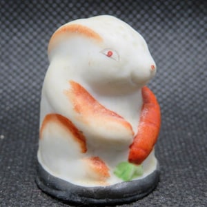 May include: A white porcelain thimble shaped like a rabbit holding a carrot. The rabbit has orange and brown markings and red eyes. The thimble has a black base.