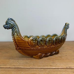 May include: A ceramic Viking ship bowl with a dragon head at the bow. The bowl is decorated with green and brown glaze and has a pattern of shields with crosses on them.
