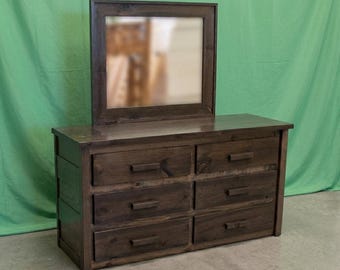 May include: A dark brown wooden dresser featuring six rectangular drawers with horizontal wooden handles. A matching rectangular mirror with a thick dark brown wooden frame is attached to the top surface of the dresser unit.