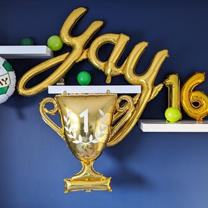 Puede incluir: Globos dorados que deletrean "yay" y "16" con un globo de fútbol verde y blanco que dice "Feliz cumpleaños" y un globo de trofeo dorado con un número "1" blanco y una corona de laurel blanca.