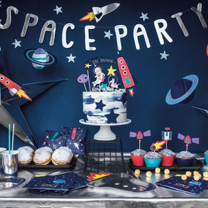 May include: A space-themed party table setting with a blue and white cake decorated with astronaut figurines, cupcakes with rocket ship toppers, and a banner that reads "Space Party".