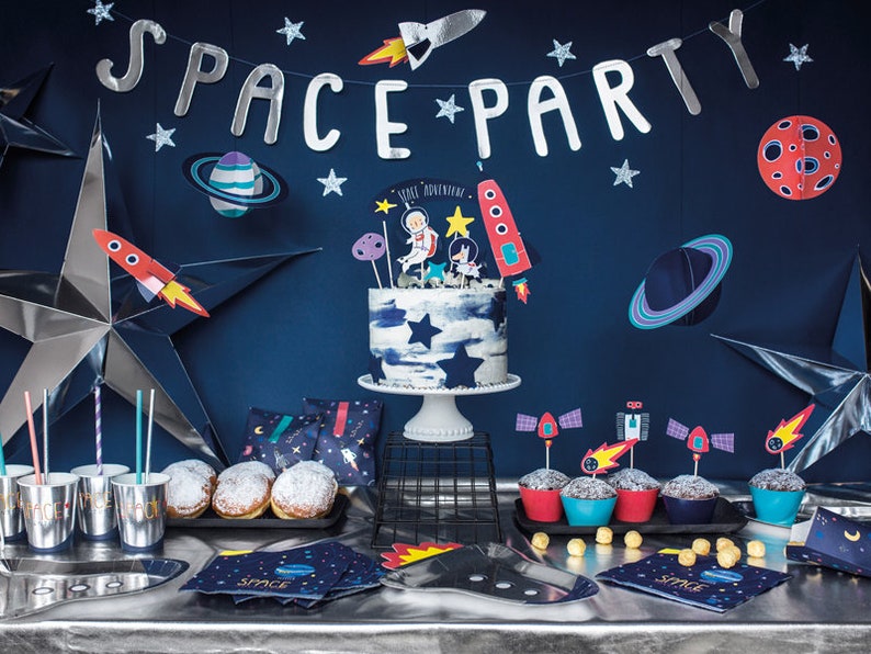 May include: A space-themed party table setting with a blue and white cake decorated with astronaut figurines, cupcakes with rocket ship toppers, and a banner that reads "Space Party".