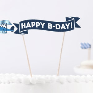 Puede incluir: Un pastel blanco con un avi&oacute;n azul y una pancarta que dice "HAPPY B-DAY!" en la parte superior.