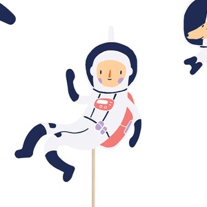 May include: A set of three astronaut-themed cake toppers. The toppers feature a white astronaut with a red and pink spacesuit, a white dog with a blue space helmet, and a blue crescent moon.