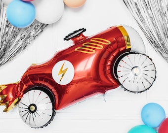 May include: A red race car-shaped balloon with gold accents and a white lightning bolt emblem. The balloon has silver and white wheels and is surrounded by colorful balloons and silver fringe.