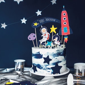 May include: A blue and white frosted cake decorated with a rocket, astronaut, and stars. The cake is on a white cake stand and has the words "Space Adventure" written on it.