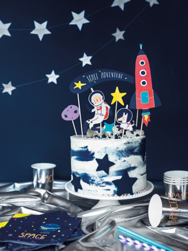 May include: A blue and white frosted cake decorated with a rocket, astronaut, and stars. The cake is on a white cake stand and has the words "Space Adventure" written on it.