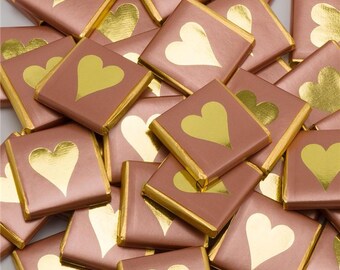 May include: A pile of small, square, pink chocolate bars with gold foil hearts. The bars are wrapped in gold foil and have a gold border.