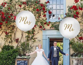 May include: A bride and groom stand in front of a building decorated with red flowers. The bride is holding a white balloon with the word "Mrs." written in gold. The groom is holding a white balloon with the word "Mr." written in gold.