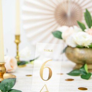 May include: Gold table number six on a clear acrylic card with a gold triangle stand. The card says "Table" at the top.