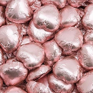Puede incluir: Primer plano de un montón de corazones de chocolate envueltos en papel de aluminio de color oro rosa. Los corazones están dispuestos en un patrón aleatorio, creando una exhibición visualmente atractiva y festiva.