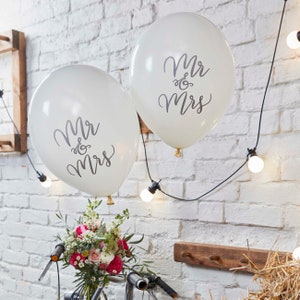 May include: Two white balloons with the text "Mr & Mrs" printed in black. The balloons are tied together and are floating in front of a white brick wall with fairy lights.