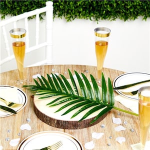 May include: A wooden slice with a green palm leaf on top, surrounded by white petals and clear glass champagne flutes with gold rims. The table is made of wood and has a white chair in the background.