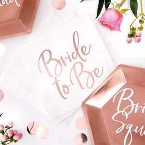 May include: Rose gold foil napkins with the text "Bride to Be" in white script.  A rose gold foil plate with the text "Bride Squad" in white script.