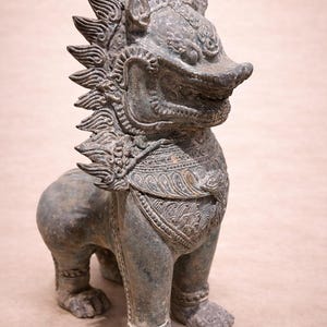 Könnte beinhalten: Verwitterte, bronzefarbene Statue einer mythischen Kreatur mit löwenartigem Körper und menschenähnlichem Gesicht. Die Statue weist aufwendige Details auf, darunter ein Kopfschmuck und eine verzierte Brustpanzerung. Die Kreatur steht.
