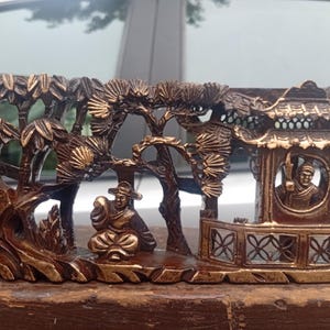 May include: A rectangular dark brown wooden carving featuring a relief scene of a figure sitting outdoors near a structure with a tiled roof, surrounded by stylised pine trees and rock formations, with gold-toned highlights on the raised surfaces.
