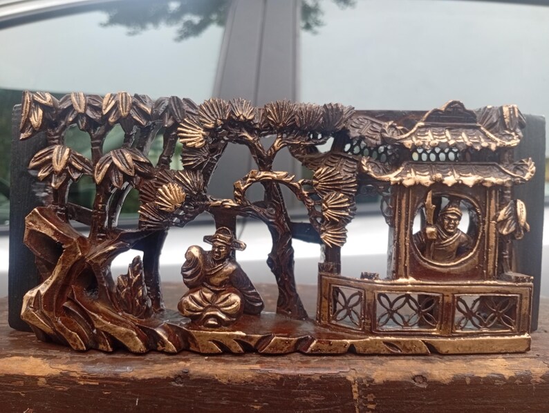 May include: A rectangular dark brown wooden carving featuring a relief scene of a figure sitting outdoors near a structure with a tiled roof, surrounded by stylised pine trees and rock formations, with gold-toned highlights on the raised surfaces.