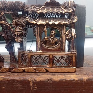 May include: A dark brown and gold-toned decorative carving featuring a detailed architectural structure with a tiled roof and a circular window. Inside the window, a figure holds a polearm. To the left, a tree with fan-shaped leaves is depicted.